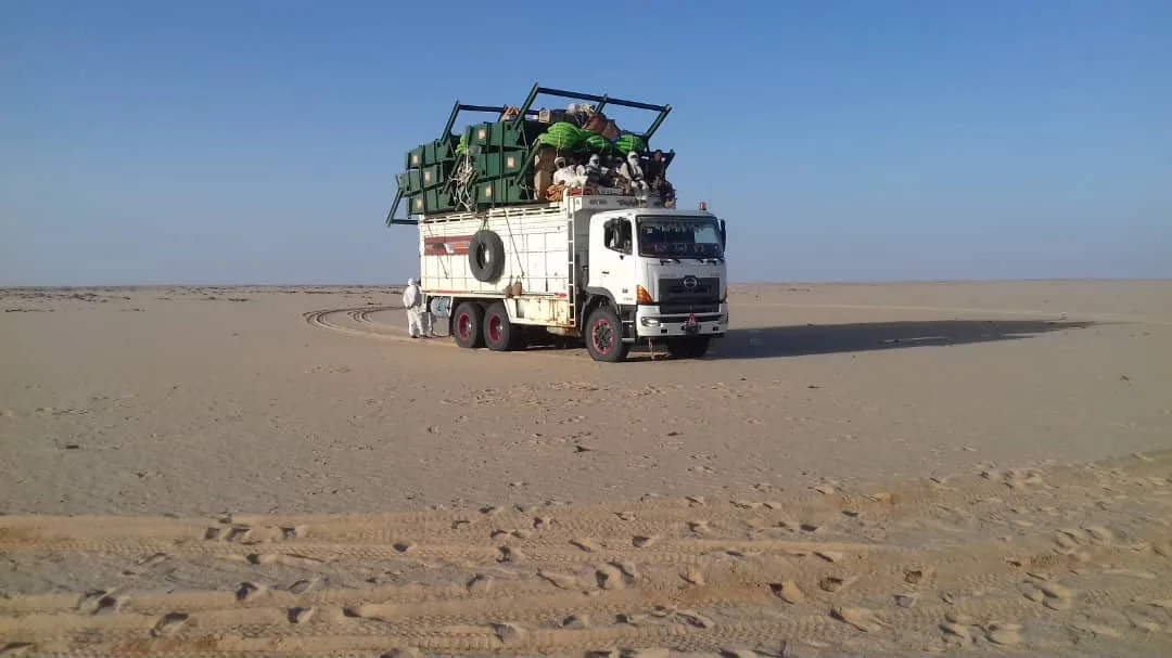 Un camion chargé de marchandises et de passagers assis sur le toit, arrêté au milieu d'une étendue de sable plate, seules des traces de pneus indiquent une piste.
